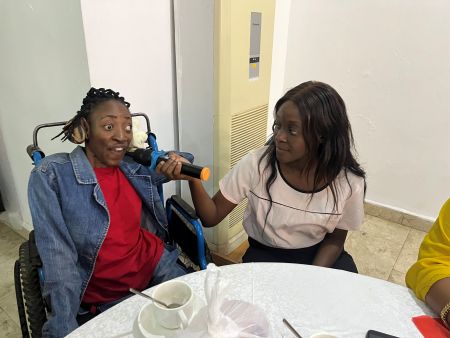 Amina, who has cerebral palsy and is using a wheelchair, is speaking into a microphone held by her personal assistant
