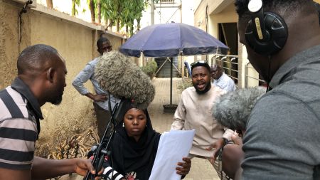 A woman in a wheelchair is reading a script. A man is acting beside her and there are microphones being held up in front of them.