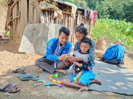 Saurab, a mobile teacher, is sitting crossed-legged on a mat with Sunmaya, a young girl with disabilities, and her mother. They are helping her to thread a green ball onto a thread of string. 
