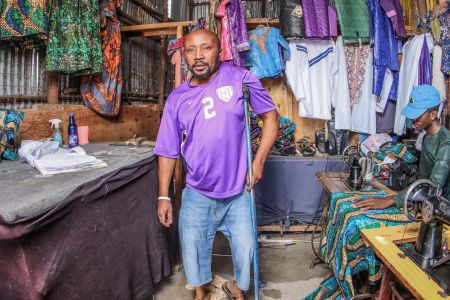 A man wearing a purple football shirt and using crutches smiles at the camera. A young man is using a sewing machine and there are different items of clothing hanging up on the walls behind him.