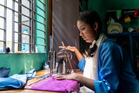 Mim, a young woman, sits opposite a window using a sewing machine. She has a physical disability which affects her hands.