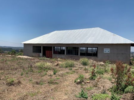 Unfinished packhouse in Lusitu Village, featuring a single-storey building with a metal roof, open window openings, and surrounding dry vegetation